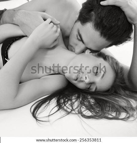 Black and white close up portrait of attractive young lovers couple together in bed with man kissing woman on neck, wearing sexy lingerie in a hotel, interior. Romance and sex lifestyle indoors.