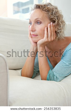 Aspirational beauty portrait of an attractive mature healthy woman laying on a white sofa at home, thoughtful and relaxing indoors. Home living and well being lifestyle, aging well middle age woman.