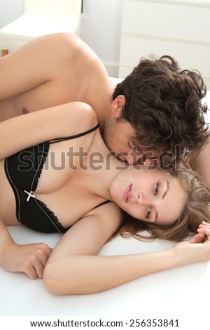 Attractive young lovers couple being together in bed with man kissing woman on neck wearing sexy lingerie in a hotel room, interior. Romance and sex lifestyle indoors. Relationships and feelings.
