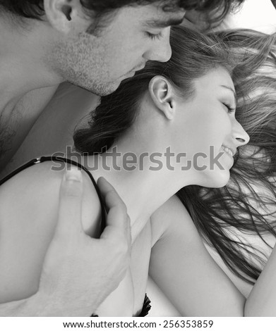 Black and white close up portrait of attractive young lovers couple together in bed with man kissing woman on neck, wearing sexy lingerie in a hotel room, interior. Romance and sex lifestyle indoors.