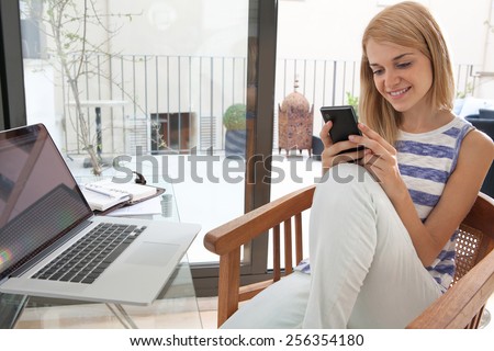 Portrait of attractive teenager girl college student sitting at her work desk at home, using a smartphone and a laptop computer to work and study. Professional young woman using mobile phone indoors.