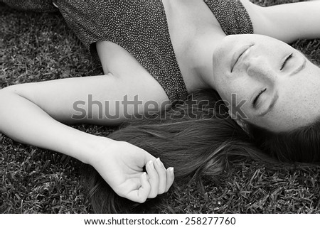 Eyes closed black and white beauty portrait of a young beautiful tourist woman laying down on grass in a park, relaxing on a summer holiday, smiling. Healthy living, beauty and well being lifestyle.