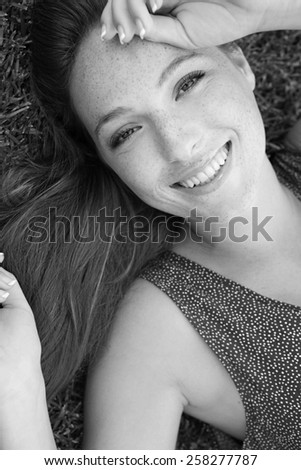 Black and white close up beauty portrait of a joyful young beautiful tourist woman laying down on grass in a park, enjoying a summer holiday, smiling. Healthy living and beauty lifestyle, exterior.