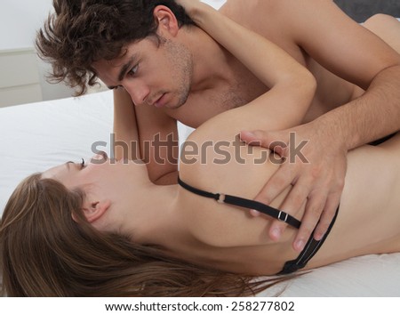 Young attractive romantic couple hugging and kissing, laying down together on a white bed, having sex and being loving with each other. Love and relationships lifestyle, interior bedroom.