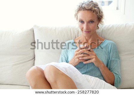 Aspirational close up beauty smiling portrait of an attractive mature healthy woman sitting down on a white sofa at home, relaxing drinking tea indoors. Home living and well being lifestyle.