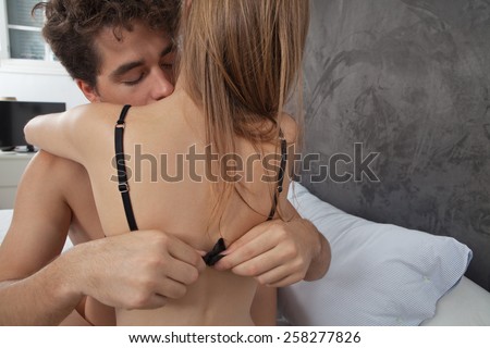 Young attractive romantic couple hugging and kissing, with man undressing woman on a white bed, having sex and being loving with each other. Love and relationships lifestyle, interior bedroom.