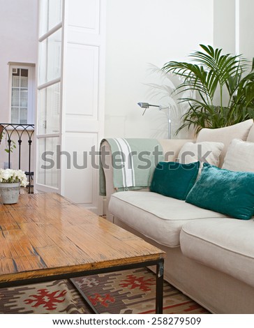 Home living room with white sofa and green cushions and plants, interior view. House indoors with carpets and picture frames, and decorative bamboo coffee table. Tranquil and aspirational home space.