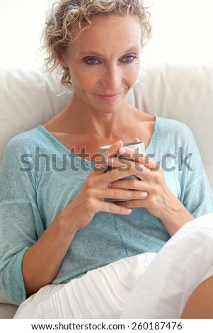 Portrait of an attractive mature caucasian woman sitting on a white sofa at home, lounging and relaxing holding a mug and drinking tea or coffee, indoors lifestyle. Health, wellness and well being.
