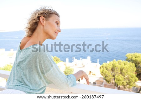 Side portrait of a mature middle age attractive woman relaxing in her holiday resort hotel balcony, contemplating the sea and blue sky during a summer vacation day, outdoors. Beauty and lifestyle.