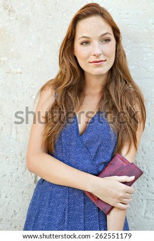Close up portrait of an elegant and smart young professional tourist woman leaning on a plain textured stone wall on holiday, carrying a purse, smiling outdoors. Travel and consumerism lifestyle.