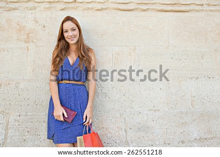 Portrait of a young professional tourist woman leaning on a plain textured stone wall on holiday, carrying shopping bags and joyfully smiling at the camera, outdoors. Travel and consumerism lifestyle.