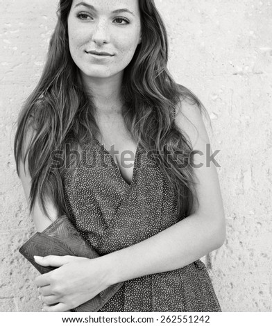 Black and white close up portrait of an elegant and smart young professional tourist woman standing leaning on a plain textured stone wall on holiday, smiling outdoors. Travel consumerism lifestyle.