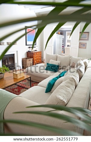 Still life of a stylish and elegant home living room with a large white sofa and a fireplace, seen though foliage green leaves, house interior. Beautiful design home space and family room, indoors.