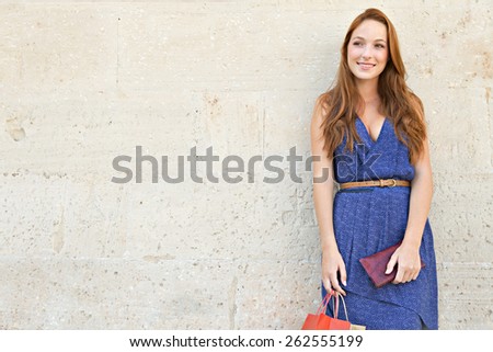 Portrait of a young professional tourist woman standing on a plain textured stone wall on holiday, with shopping bags and a purse, joyfully smiling, outdoors. Travel and consumerism lifestyle.