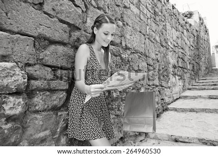 Black and white portrait of young tourist woman visiting a destination city with old textured stone wall in a street with steps, using a map, sightseeing on holiday. Travel vacation, outdoors.