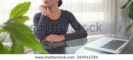 Panoramic faceless view of a young professional business woman sitting at her office space work desk being thoughtful, with laptop computer and smart phone device, against a bright window.