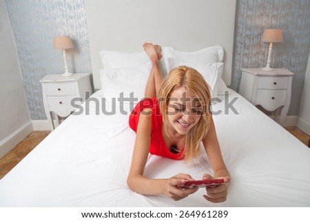 Attractive young teenager woman laying down and relaxing in a stylish decorative home bedroom using a smartphone device to network and go on line. Home interior technology lifestyle, indoors.