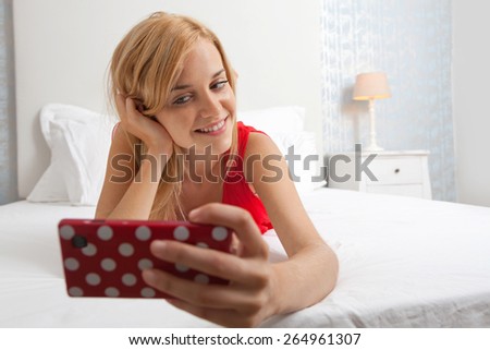 Portrait of attractive young teenager woman laying down in stylish home bedroom using a smartphone device to take selfies pictures of herself, networking. Home interior technology lifestyle, indoors.