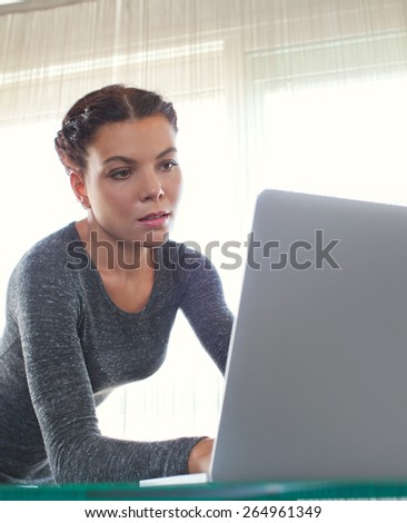 Young attractive professional business woman at office space work desk reaching and typing on her laptop computer against a bright window, working. Business finance and administrative job, interior.