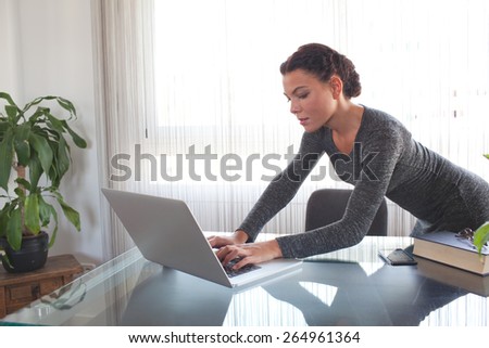 Young attractive professional business woman at office space work desk reaching and typing on her laptop computer against a bright window with reflections on glass table. Business finance, interior.