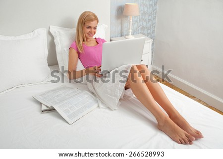 Beautiful teenager woman relaxing on a bed in a home bedroom using a laptop computer to shop on line, smiling house interior. Connectivity technology buying and spending lifestyle, hotel interior.