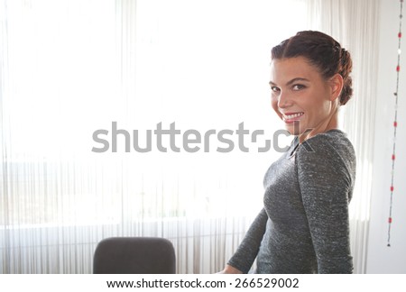 Portrait of young professional business woman sitting at her office space work desk, turning to smile at the camera joyful against a sunny window, indoors. Business finance office interior lifestyle.