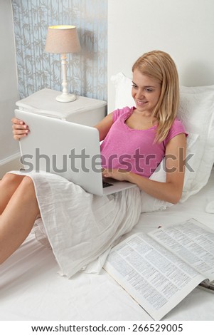 Beautiful teenager woman relaxing on bed in a home bedroom using a laptop computer to shop on line, with magazines and smiling, house interior. Connectivity technology lifestyle, hotel room interior.