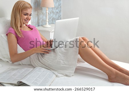 Beautiful teenager woman relaxing on bed in a home bedroom using a laptop computer to shop on line, with magazines and smiling, house interior. Connectivity technology lifestyle, hotel room interior.