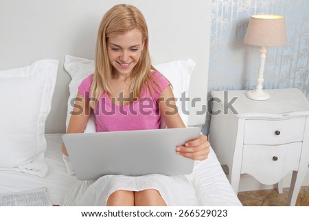 Portrait of beautiful teenager woman relaxing on a bed in a bedroom using a laptop computer to study, with books and smiling, house interior. Connectivity technology student lifestyle, home interior.