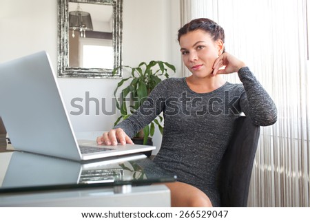 Portrait of an attractive young office business woman sitting smiling in her home office desk, workplace interior. Professional working woman using technology at work, indoors.
