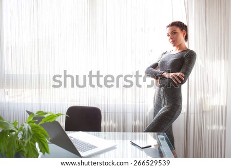 Side view of office business woman standing with arms crossed by bright window in home office desk, thoughtful and contemplative, interior. Aspirational professional woman using technology, indoors.