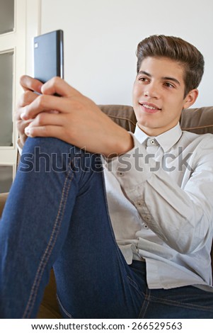 Portrait of a fashionable teenager young man wearing a shirt and lounging on a sofa at home, using a smart phone for networking on line, home interior. Young modern man smiling, lifestyle indoors.