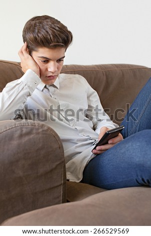 Portrait of fashionable teenager young man wearing a shirt lounging on sofa at home, holding a digital smart phone for networking on line, home interior. Young thoughtful modern man lifestyle indoors.