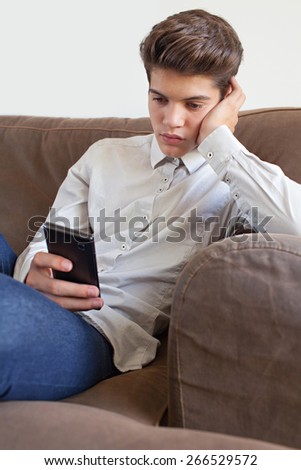 Portrait of fashionable teenager young man wearing a shirt lounging on a sofa at home, using a smart phone device for networking on line, home interior. Young thoughtful modern man lifestyle indoors.