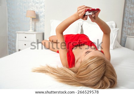 Over head view of attractive young woman laying relaxing in a stylish home bedroom using a smartphone device touch screen to network on line. Home interior connectivity technology lifestyle, indoors.