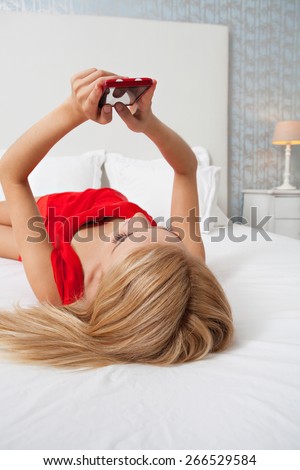 Attractive young teenager woman laying relaxing in stylish decorative home bedroom using a smartphone device touch screen to network on line. Home interior connectivity technology lifestyle, indoors.