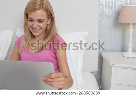Portrait of beautiful teenager woman relaxing on a bed in a bedroom using a laptop computer using technology and the internet, smiling indoors. Connectivity technology lifestyle, room interior.