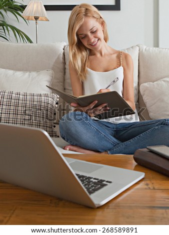 Attractive young woman sitting on sofa at home, using laptop technology and telecommunications, writing on a notepad smiling. Student girl doing homework at home, interior. Home technology lifestyle.