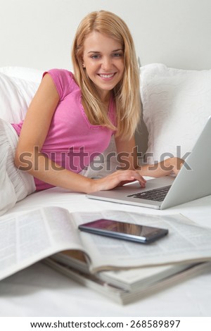 Portrait of attractive young teenager woman laying down in stylish decorative white home bedroom using laptop computer and joyfully smiling at the camera. Home interior technology lifestyle, indoors.
