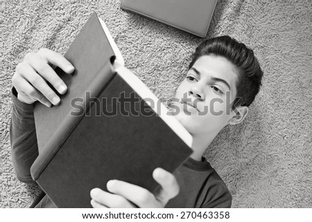 Black and white close up portrait of a young student man reading a book laying down on bed at home. Boy studying at home laying on a carpet doing his homework and learning indoors. Student home life.