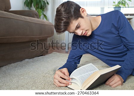 Portrait of attractive teenager man holding a text book open while laying down on a rug in a home living room with a sofa, reading and studying at home, interior. Student living, interior lifestyle.