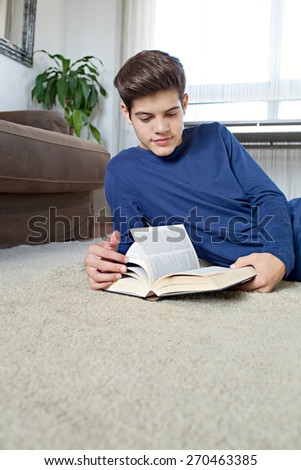 Portrait of an attractive teenager man holding a text book open while laying down on a rug in home living room with a sofa, reading and studying at home, interior. Student living, interior lifestyle.