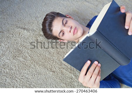 Portrait of a young student man holding a notebook and reading while laying down on a bed at home. Boy studying at home laying on a carpet doing his homework and learning indoors. Student home life.