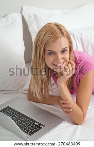 Portrait of attractive teenager woman relaxing in a stylish white home bedroom using a laptop computer and joyfully smiling at the camera, house interior. Home interior technology lifestyle, indoors.