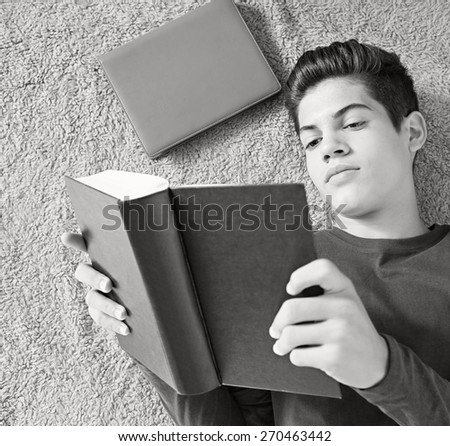 Black and white close up portrait of a young student man reading a book laying down on bed at home. Boy studying at home laying on a carpet doing his homework and learning indoors. Student home life.