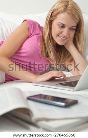 Portrait of attractive woman relaxing in stylish decorative white home bedroom using a laptop computer, with magazines and a smartphone smiling, interior. Home interior technology lifestyle, indoors.