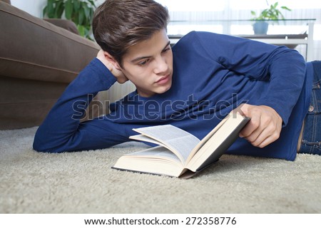 Portrait of a student teenager man with open book laying down on a rug in home living room with sofa, reading and studying at home, interior. Student boy in house living, interior lifestyle.
