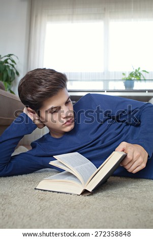 Portrait of a student teenager holding a text book open laying down on a rug in a home living room with sofa, reading and studying at home, interior. Student boy in house living, interior lifestyle.