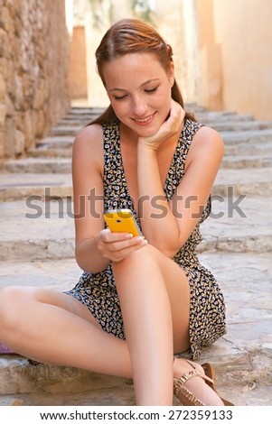 Portrait of a young attractive tourist woman sitting on the stone steps of a destination city street on a sunny day on vacation, using a smartphone to network on line, smiling. Travel and technology.