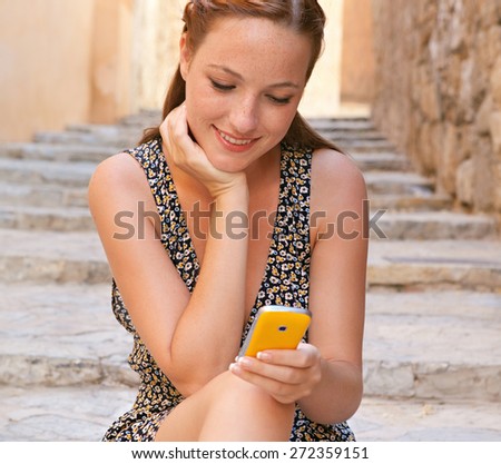 Portrait of a young attractive tourist woman sitting on the stone steps of a destination city street on a sunny day on vacation, using a smartphone to network on line, smiling. Travel and technology.